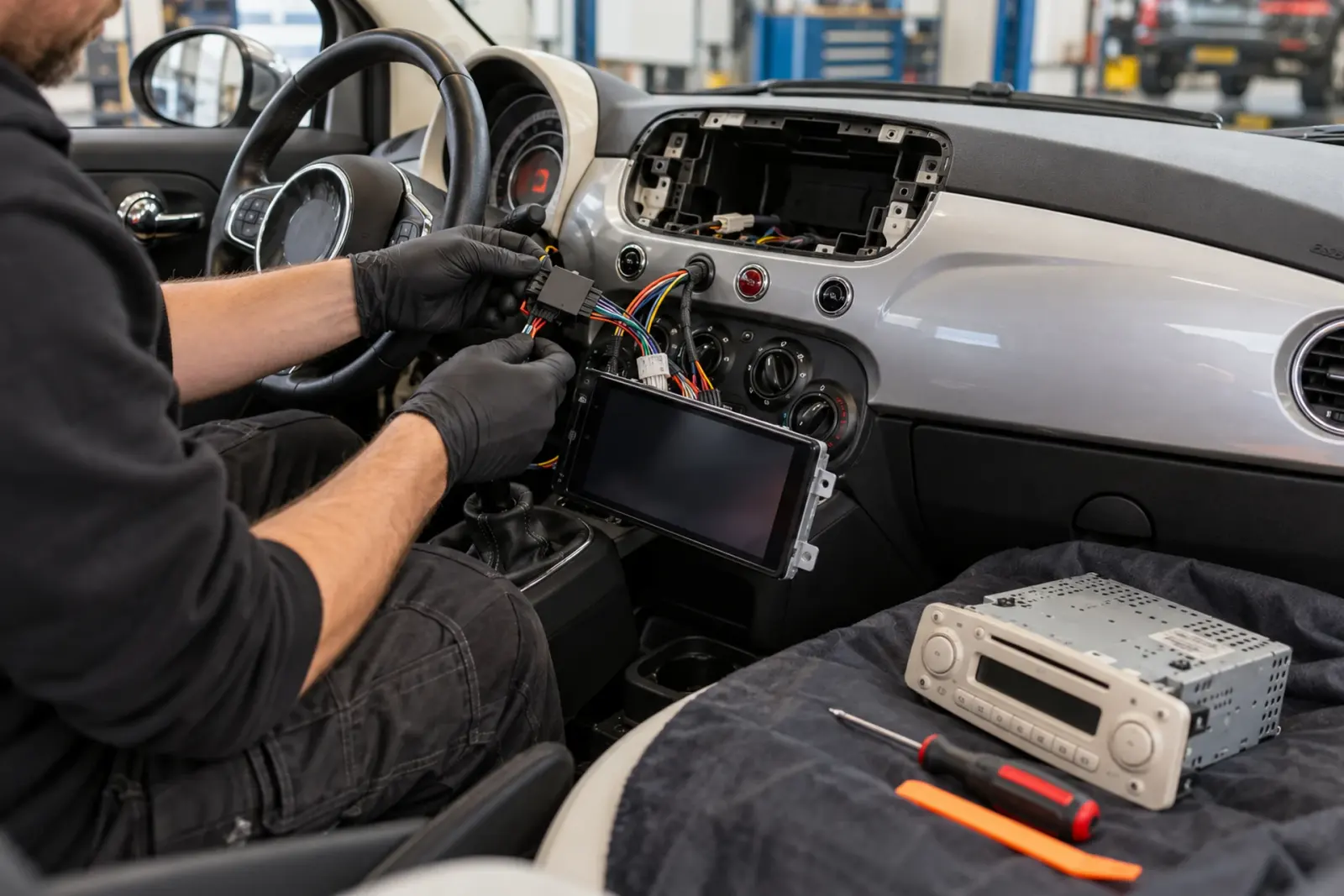 Android Auto radio inbouwen in 's-Hertogenbosch: monteur met handschoenen sluit de kabelboom van een nieuwe touchscreen-radio aan in een Fiat 500, de oude fabrieksradio ligt op de stoel